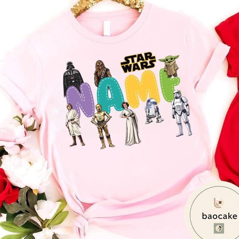 May include: A light pink t-shirt featuring Star Wars characters and the word "NAME" in colorful letters. Characters include Darth Vader, Chewbacca, Yoda, and a Stormtrooper. The Star Wars logo is also visible.