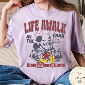 May include: A light purple t-shirt featuring a graphic of Mickey Mouse in front of a castle with the text "LIFE AWALK IN THE PARK" and "Walt Disney World" in red and white. The shirt has short sleeves.