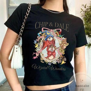 May include: Black t-shirt featuring Chip and Dale in wizard attire, surrounded by a floral wreath. The text "Chip & Dale" arches across the top, with "Wizard Wonders" below. The design includes a blue wizard hat and red robes.