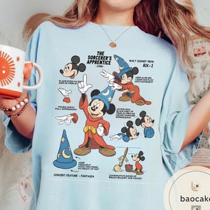 May include: Light blue t-shirt featuring a graphic of Mickey Mouse as the Sorcerer's Apprentice, with text and illustrations from the 1940 Disney film. The design includes a blue wizard hat with a crescent moon and stars.