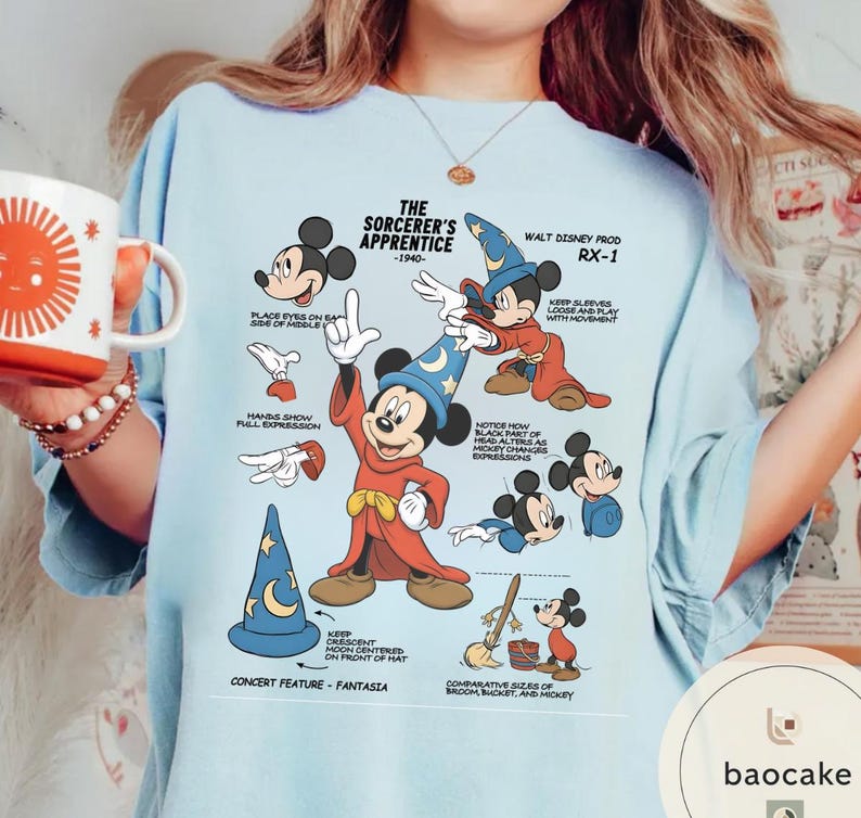 May include: Light blue t-shirt featuring a graphic of Mickey Mouse as the Sorcerer's Apprentice, with text and illustrations from the 1940 Disney film. The design includes a blue wizard hat with a crescent moon and stars.