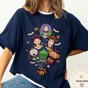Pode incluir: Uma t-shirt azul-marinho com uma estampa gr&aacute;fica de v&aacute;rios personagens animados vestidos com fantasias de Halloween, incluindo um patrulheiro espacial, uma cowgirl, um cowboy, um dinossauro, um c&atilde;o e uma batata, presos a um bal&atilde;o de ab&oacute;bora.