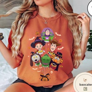Pode incluir: Uma camisola laranja de gola redonda com uma impress&atilde;o gr&aacute;fica de v&aacute;rias personagens animadas vestidas com fatos de Halloween, incluindo um ranger espacial, uma cowgirl, um xerife, um c&atilde;o, um dinossauro e uma figura de batata, presos a um bal&atilde;o de ab&oacute;bora.