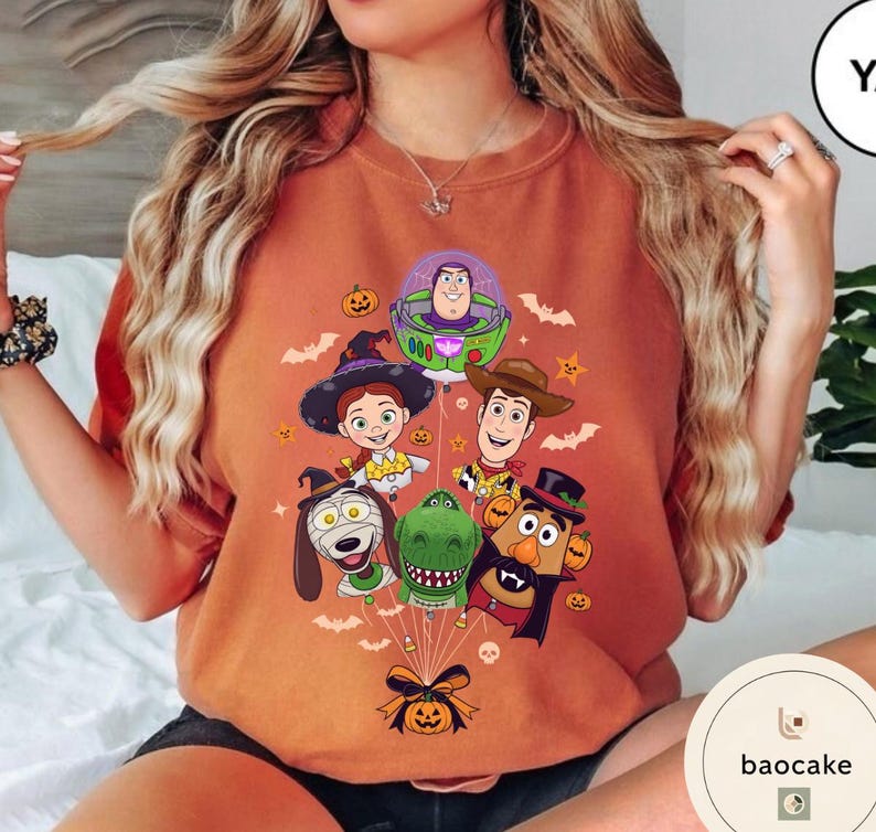 Pode incluir: Uma camisola laranja de gola redonda com uma impress&atilde;o gr&aacute;fica de v&aacute;rias personagens animadas vestidas com fatos de Halloween, incluindo um ranger espacial, uma cowgirl, um xerife, um c&atilde;o, um dinossauro e uma figura de batata, presos a um bal&atilde;o de ab&oacute;bora.