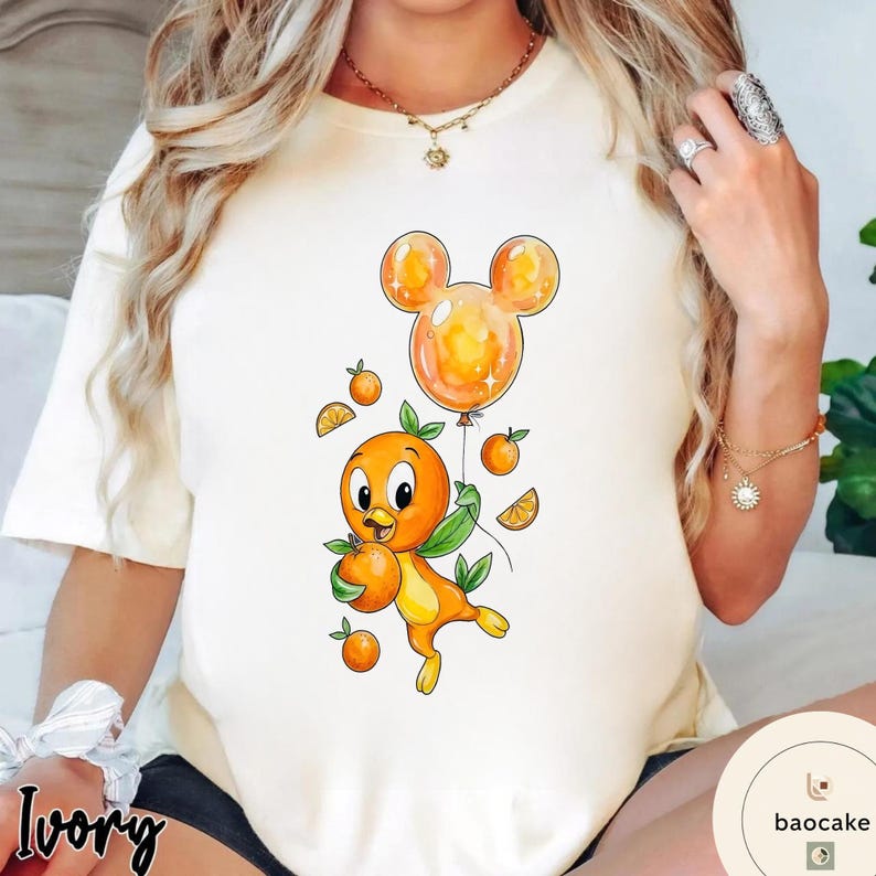 Op de afbeelding: Ivoor kleurig T-shirt met een cartoon oranje vogel die sinaasappels en een Mickey Mouse-vormige ballon vasthoudt. De vogel heeft groene bladeren en is omringd door sinaasappelschijfjes en kleine sinaasappels. Het shirt heeft een ronde hals.