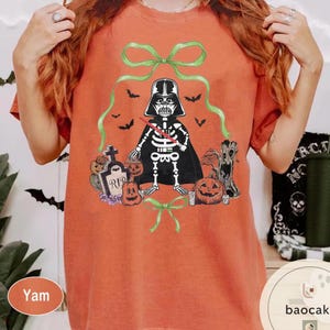 May include: Orange t-shirt featuring a Halloween-themed graphic. The design includes a skeleton dressed as Darth Vader, surrounded by pumpkins, bats, and a tombstone. Green bows and a cape complete the spooky look.