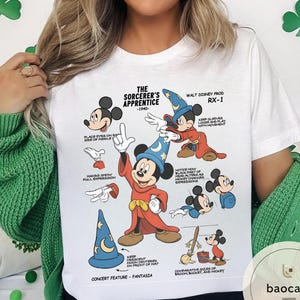 May include: White t-shirt featuring a graphic of Mickey Mouse as the Sorcerer's Apprentice. The design includes illustrations and text detailing the character's features and the film's production details. The shirt has a crew neck and short sleeves.