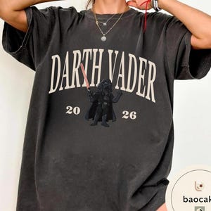 May include: Dark gray t-shirt featuring the text "DARTH VADER" in white, with a graphic of Darth Vader holding a red lightsaber. The numbers "20" and "26" are below the text and graphic. The shirt has a vintage, worn look.