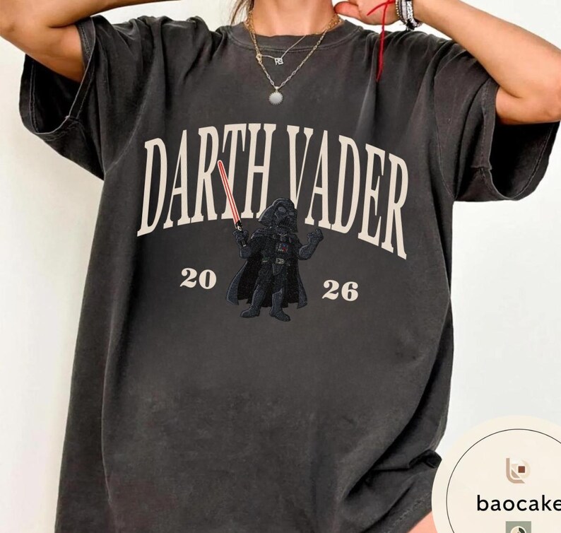 May include: Dark gray t-shirt featuring the text "DARTH VADER" in white, with a graphic of Darth Vader holding a red lightsaber. The numbers "20" and "26" are below the text and graphic. The shirt has a vintage, worn look.