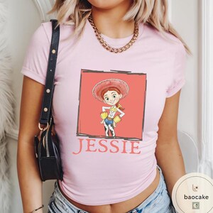 K&ouml;nnte beinhalten: Ein hellrosa T-Shirt mit kurzen &Auml;rmeln und einer Grafik der Figur Jessie aus Toy Story in einem korallenfarbenen Quadrat. Der Name JESSIE ist in Rot unter der Grafik aufgedruckt. Eine goldfarbene Kette wird zu dem T-Shirt getragen.