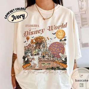 Op de afbeelding: Ivoor kleurig T-shirt met een Halloween-thema grafische afbeelding met de tekst "Florida Disney World" en griezelige illustraties. Het ontwerp bevat een mummie, pompoenen, vleermuizen en een pompoen. De tekst "The Most Scariest Place On Earth" staat ook op het shirt.