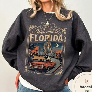 May include: Dark gray sweatshirt featuring a graphic of Mickey Mouse driving a red convertible towards a castle with the text "Welcome to Florida" and "Est. 1971". The design includes palm trees and a crescent moon.