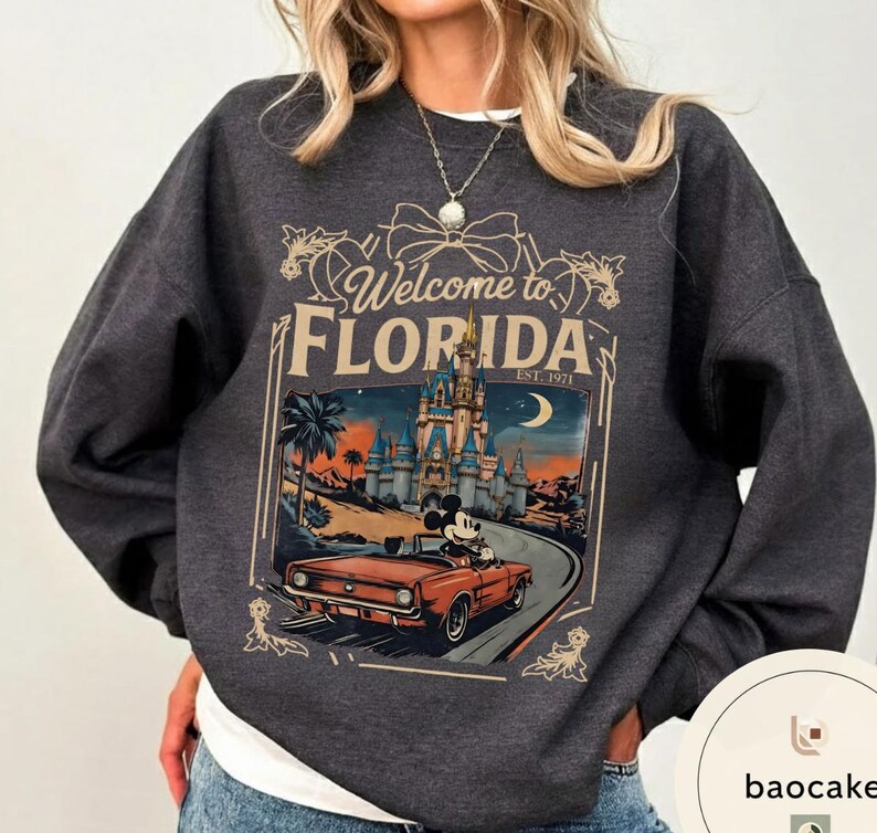 May include: Dark gray sweatshirt featuring a graphic of Mickey Mouse driving a red convertible towards a castle with the text "Welcome to Florida" and "Est. 1971". The design includes palm trees and a crescent moon.