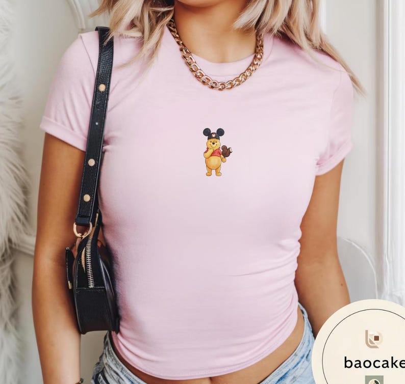May include: A light pink short-sleeved t-shirt featuring a small embroidered graphic of a yellow bear wearing black mouse ears and holding a brown object. The shirt has a crew neckline and is paired with a black shoulder bag and a gold chain necklace.