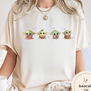 May include: Cream-colored t-shirt featuring four cartoon illustrations of a green alien character. Each character is in a different pose, with various outfits and accessories. The design includes small stars and sparkles. The brand name "baocake" is printed on the bottom right.