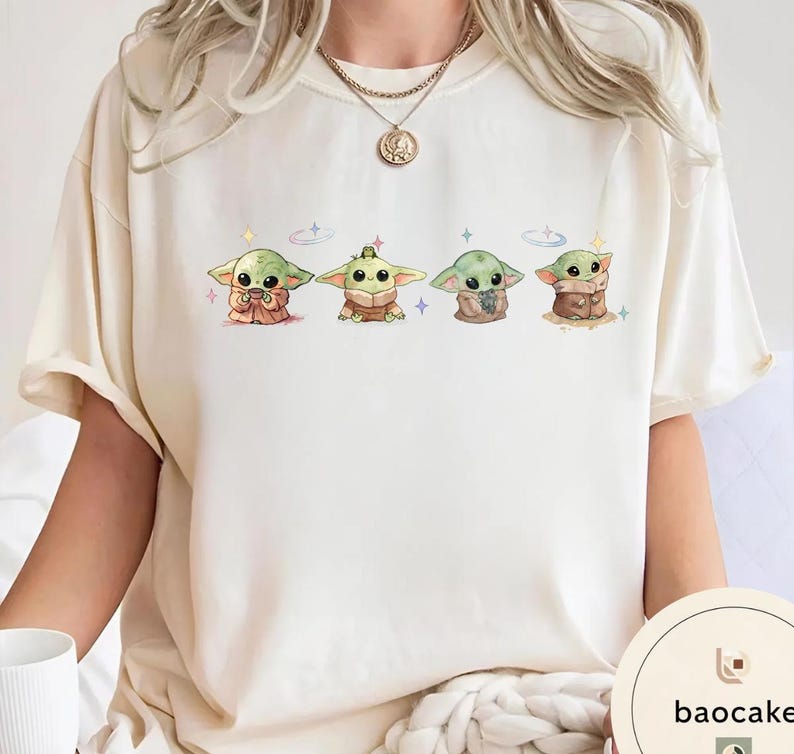 May include: Cream-colored t-shirt featuring four cartoon illustrations of a green alien character. Each character is in a different pose, with various outfits and accessories. The design includes small stars and sparkles. The brand name "baocake" is printed on the bottom right.