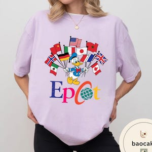May include: A lilac t-shirt with a graphic of Donald Duck holding flags from different nations. The word "Epcot" is written in colourful letters below the character, with a globe graphic in the letter "C".