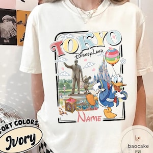 Puede incluir: Camiseta de color marfil con un gr&aacute;fico colorido de Tokyo Disneyland. El dise&ntilde;o incluye las palabras "TOKYO Disneyland", una estatua, Mickey Mouse, el Pato Donald, un castillo y un globo de arco&iacute;ris. La camiseta tambi&eacute;n tiene la palabra "Name".