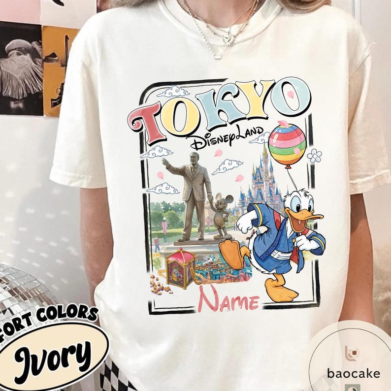 Puede incluir: Camiseta de color marfil con un gr&aacute;fico colorido de Tokyo Disneyland. El dise&ntilde;o incluye las palabras "TOKYO Disneyland", una estatua, Mickey Mouse, el Pato Donald, un castillo y un globo de arco&iacute;ris. La camiseta tambi&eacute;n tiene la palabra "Name".