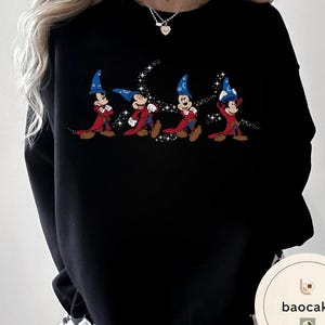 May include: Black sweatshirt featuring a cartoon graphic of Mickey Mouse in a sorcerer's outfit. The design shows Mickey in a red robe and blue wizard hat, with a trail of sparkling stars. The sweatshirt is a casual style.