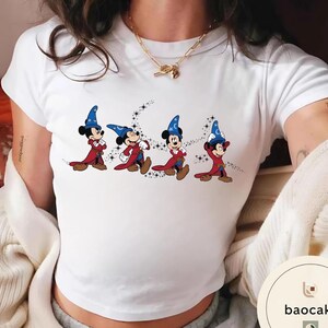 May include: White t-shirt featuring a cartoon illustration of Mickey Mouse in a wizard costume. The design shows Mickey Mouse in a red robe and blue wizard hat, with a crescent moon and stars background. The shirt is a crew neck.