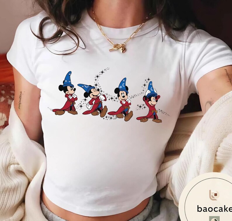 May include: White t-shirt featuring a cartoon illustration of Mickey Mouse in a wizard costume. The design shows Mickey Mouse in a red robe and blue wizard hat, with a crescent moon and stars background. The shirt is a crew neck.