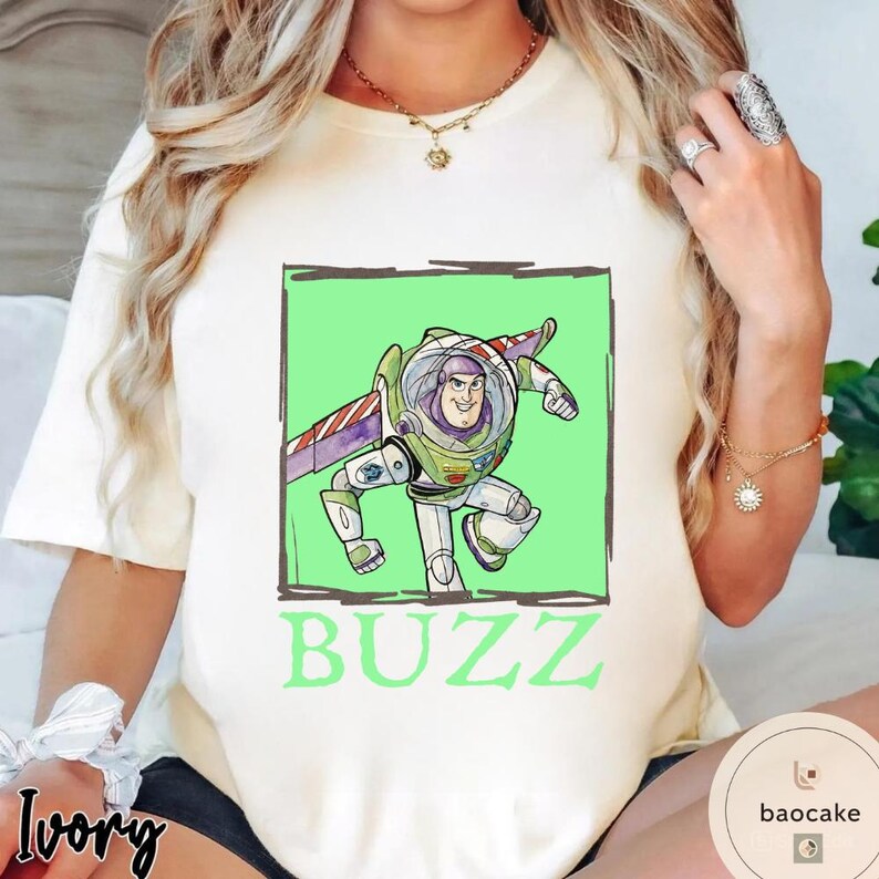 K&ouml;nnte beinhalten: Ein elfenbeinfarbenes Kurzarm-T-Shirt mit einer Grafik der Figur Buzz Lightyear in einem hellgr&uuml;nen quadratischen Rahmen. Die Figur tr&auml;gt einen wei&szlig;en, gr&uuml;nen und violetten Raumanzug mit Fl&uuml;geln. Das Wort BUZZ ist darunter in Gr&uuml;n aufgedruckt.