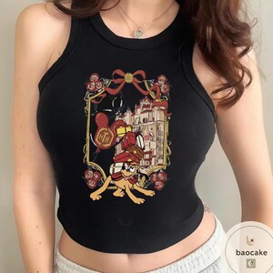 May include: Black tank top with a colorful graphic featuring a cartoon dog carrying luggage. The graphic includes a building, balloons, and the text "HTH" and "VERSE". The design is framed with roses and a bow.