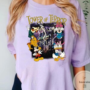 May include: A light purple t-shirt featuring a graphic of Disney characters, including Mickey Mouse, Minnie Mouse, Goofy, and Donald Duck, with the text "Tower of Terror". The graphic has a vintage, cartoon style.