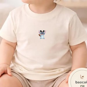 May include: A cream-colored short-sleeve t-shirt featuring a small, centered graphic of a blue alien character wearing black mouse ears and holding a brown object. The shirt has a round neckline and a simple, plain design on the front.