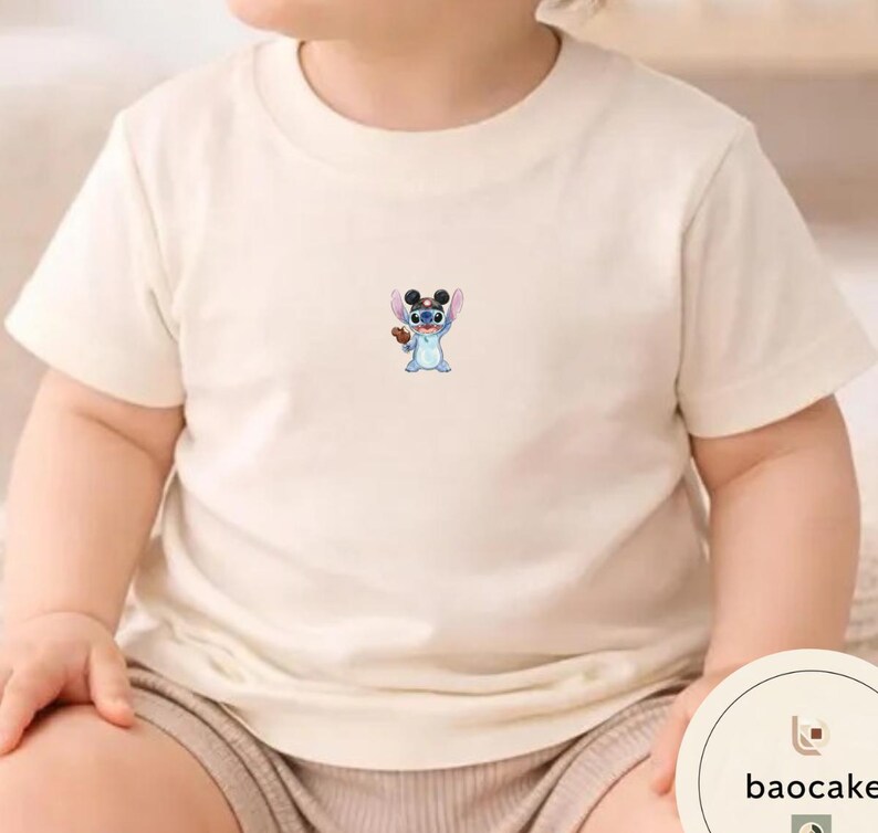 May include: A cream-colored short-sleeve t-shirt featuring a small, centered graphic of a blue alien character wearing black mouse ears and holding a brown object. The shirt has a round neckline and a simple, plain design on the front.