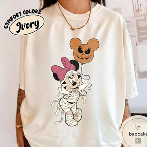 May include: Ivory Comfort Colors t-shirt featuring a Halloween-themed Minnie Mouse design. Minnie is dressed as a mummy and holding a Jack Skellington-style balloon. The shirt has the words "Comfort Colors" and "Ivory" printed on it.