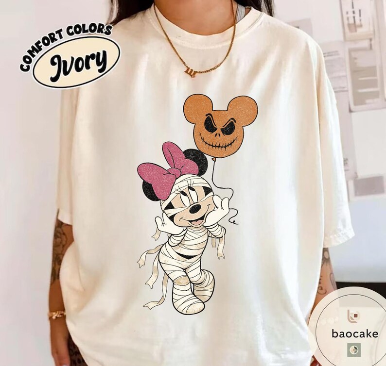 May include: Ivory Comfort Colors t-shirt featuring a Halloween-themed Minnie Mouse design. Minnie is dressed as a mummy and holding a Jack Skellington-style balloon. The shirt has the words "Comfort Colors" and "Ivory" printed on it.