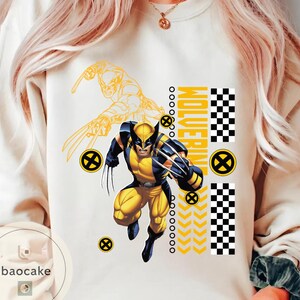 May include: Cream-colored sweatshirt featuring a graphic of Wolverine in a yellow and blue costume with claws extended. The word "Wolverine" is printed vertically in yellow, with additional yellow and black design elements.