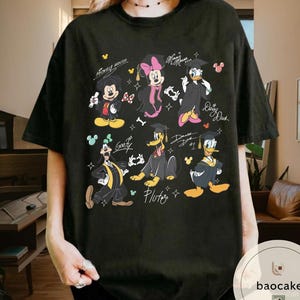 May include: Black t-shirt featuring a graphic of Mickey Mouse, Minnie Mouse, Donald Duck, Daisy Duck, Goofy, and Pluto in graduation attire. The design includes the characters' names and graduation year. The shirt has a crew neck and short sleeves.