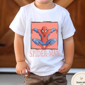 May include: White t-shirt featuring a Spider-Man graphic. The design shows Spider-Man in a crouching pose within a black outlined square on a coral background. The words "SPIDER-MAN" are printed below the graphic in coral.