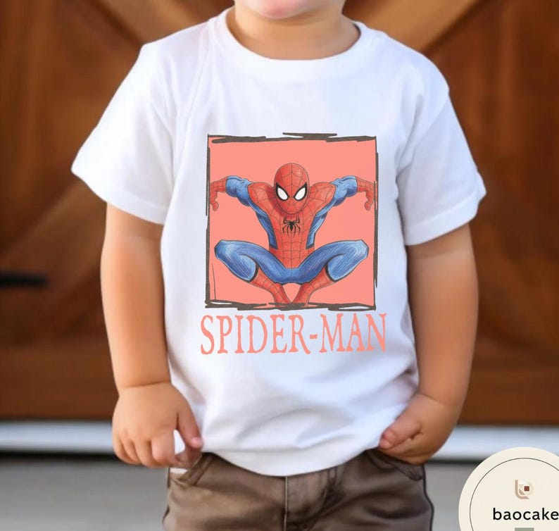 May include: White t-shirt featuring a Spider-Man graphic. The design shows Spider-Man in a crouching pose within a black outlined square on a coral background. The words "SPIDER-MAN" are printed below the graphic in coral.