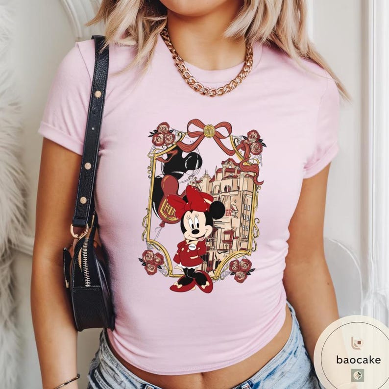 May include: A light pink t-shirt featuring a Minnie Mouse graphic. The graphic includes Minnie Mouse in a red dress and bow, a castle, and the word "Paris" in a gold frame. The shirt has short sleeves.