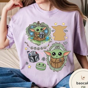 May include: A light purple t-shirt featuring a graphic print of the character Grogu in various poses, including holding a helmet, alongside a Mandalorian helmet, a green macaron, small stars, and the name Grogu spelled out on a banner.