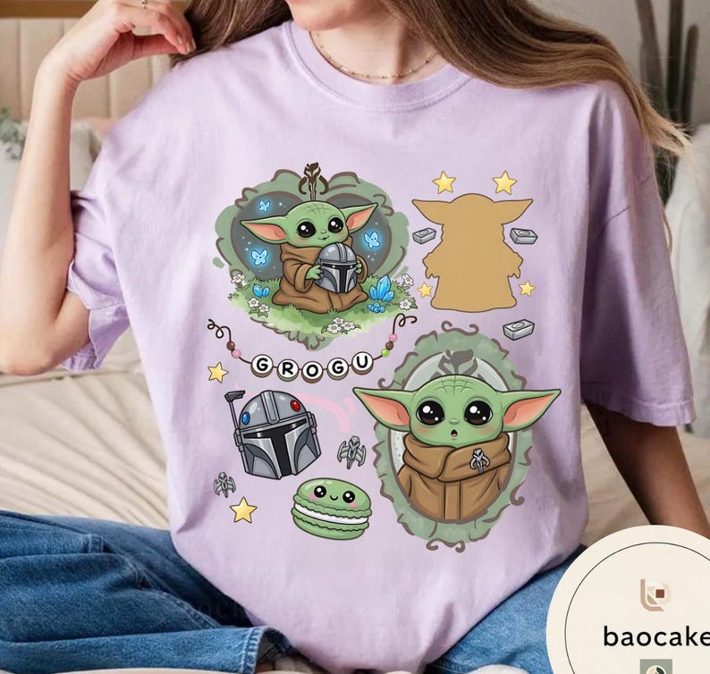 May include: A light purple t-shirt featuring a graphic print of the character Grogu in various poses, including holding a helmet, alongside a Mandalorian helmet, a green macaron, small stars, and the name Grogu spelled out on a banner.