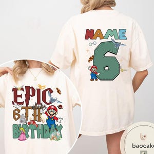May include: Cream-colored t-shirt with a colorful graphic design for a 6th birthday. The front features the text "EPIC 6TH BIRTHDAY" with cartoon characters. The back has a large number 6 and the word "NAME" above it, with cartoon characters.