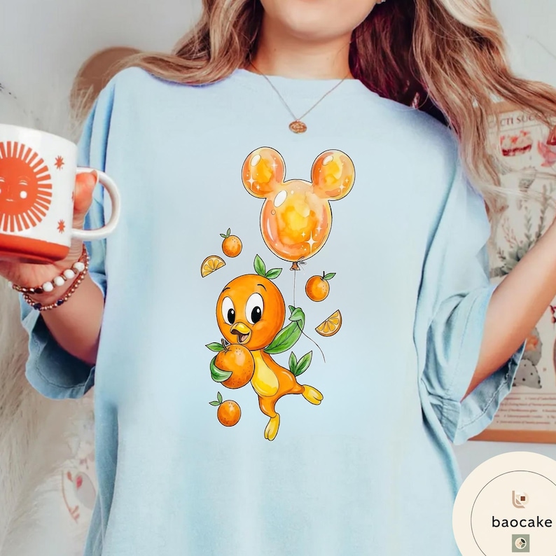 Op de afbeelding: Lichtblauw T-shirt met een cartoon oranje vogel die sinaasappels en een Mickey Mouse-vormige ballon vasthoudt. De vogel heeft groene bladeren en is omringd door sinaasappelschijfjes en hele sinaasappels. Het ontwerp is grillig en kleurrijk.