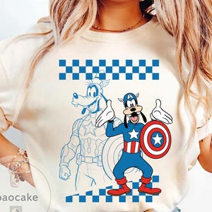 May include: Cream-colored t-shirt featuring a cartoon Goofy character dressed as Captain America, holding a shield. A blue outline of Goofy in a Captain America suit is behind him. The shirt has a blue and white checkered pattern at the top and bottom.