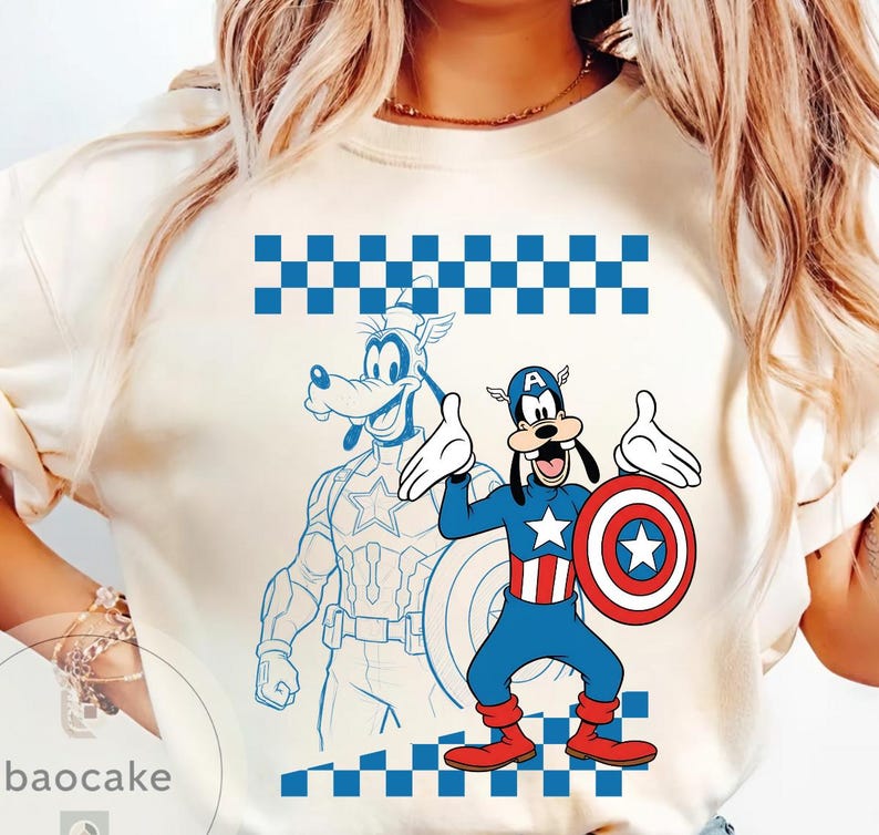 May include: Cream-colored t-shirt featuring a cartoon Goofy character dressed as Captain America, holding a shield. A blue outline of Goofy in a Captain America suit is behind him. The shirt has a blue and white checkered pattern at the top and bottom.