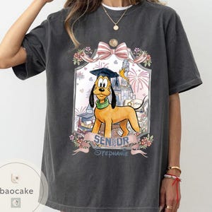 May include: Dark gray t-shirt featuring Pluto the dog wearing a graduation cap, with the text "Senior Stephanie" on a banner. The design includes a castle, fireworks, and floral accents, creating a celebratory theme.