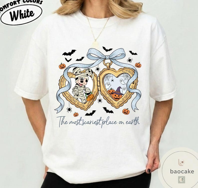 May include: White t-shirt with a Halloween design. The graphic shows a heart-shaped locket with Mickey Mouse as a mummy and Halloween elements. The text "The most scariest place on earth" is below the locket.