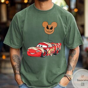 May include: Olive green t-shirt featuring a graphic of a red race car wrapped in bandages, with the number 95 on the side. A brown balloon with a jack-o'-lantern face floats above the car. The shirt has a crew neck and short sleeves.