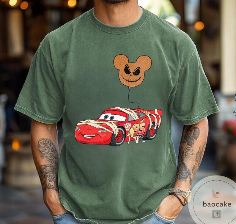 May include: Olive green t-shirt featuring a graphic of a red race car wrapped in bandages, with the number 95 on the side. A brown balloon with a jack-o'-lantern face floats above the car. The shirt has a crew neck and short sleeves.