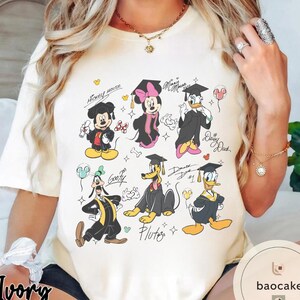 May include: Ivory-colored t-shirt featuring cartoon characters Mickey Mouse, Minnie Mouse, Donald Duck, Daisy Duck, Goofy, and Pluto in graduation attire. The design includes graduation caps, gowns, and celebratory elements.