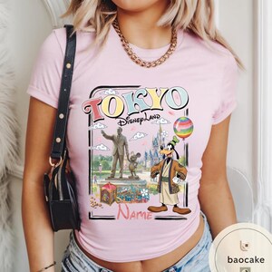 Puede incluir: Camiseta rosa claro con un dise&ntilde;o de Tokyo Disneyland. El dise&ntilde;o incluye el texto "TOKYO Disneyland" en una fuente colorida, junto con ilustraciones de Goofy, Mickey Mouse y un castillo. Tambi&eacute;n se incluye un globo de arco&iacute;ris.